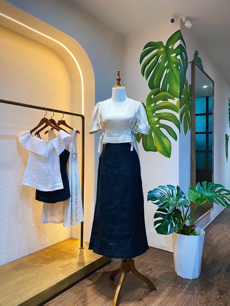 Puede incluir: Una blusa blanca de lino con mangas cortas y una falda negra de lino. La blusa tiene un detalle de lazo en la cintura. La falda tiene una cintura alta y un corte holgado. El atuendo se muestra en un maniqu&iacute; en un entorno de tienda.