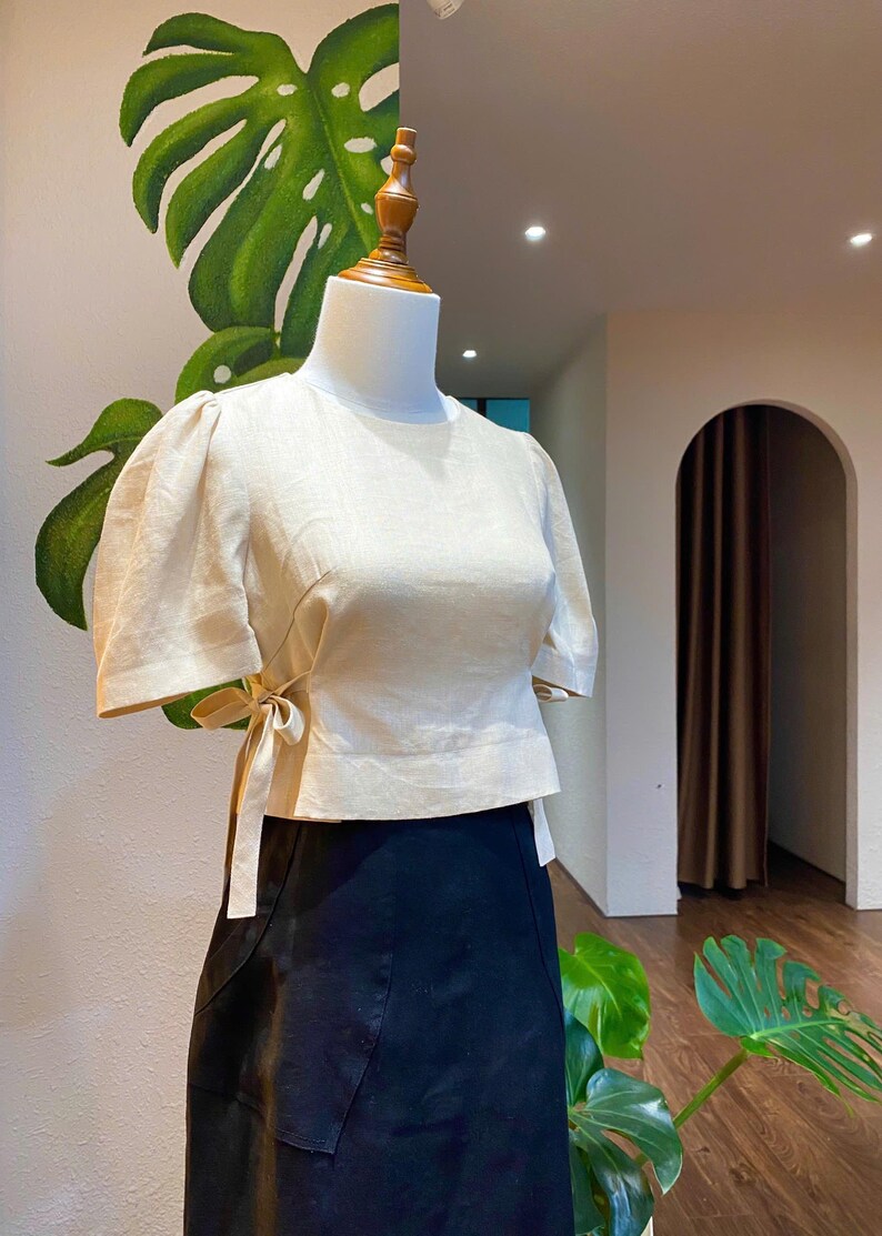 Puede incluir: Una blusa de lino beige con mangas cortas abullonadas y un lazo en la cintura. La blusa est&aacute; en un maniqu&iacute; y se combina con una falda negra.