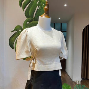 Puede incluir: Una blusa de lino beige con mangas abullonadas cortas y un lazo en la cintura. La blusa está en un maniquí blanco sobre un fondo verde y blanco.