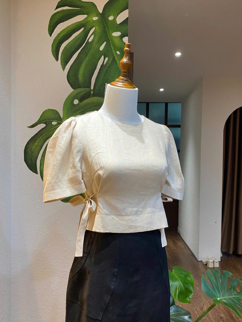Puede incluir: Una blusa de lino beige con mangas abullonadas cortas y un lazo en la cintura. La blusa est&aacute; en un maniqu&iacute; blanco sobre un fondo verde y blanco.