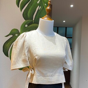 Puede incluir: Una blusa de lino color crema con mangas abullonadas cortas y un lazo en la cintura.