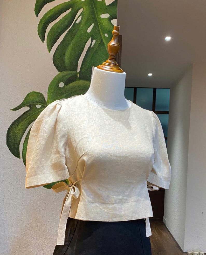 Puede incluir: Una blusa de lino color crema con mangas abullonadas cortas y un lazo en la cintura.