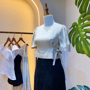 Puede incluir: Una blusa blanca de lino con mangas abullonadas cortas y un detalle de lazo en la cintura. La blusa se combina con una falda negra.