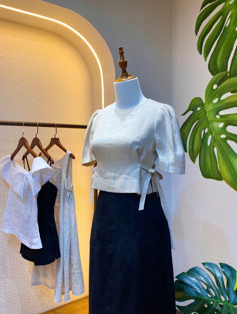 Puede incluir: Una blusa blanca de lino con mangas abullonadas cortas y un detalle de lazo en la cintura. La blusa se combina con una falda negra.