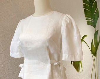 May include: A white linen top with puffed sleeves and a tie at the waist. The top is paired with matching white linen shorts.