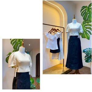 Puede incluir: Dos blusas blancas de lino con mangas abullonadas y un detalle de lazo en la cintura. Una blusa est&aacute; en un maniqu&iacute; y la otra est&aacute; colgada en una percha. Ambas blusas se combinan con faldas negras.