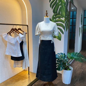 Puede incluir: Una blusa de lino blanca con mangas abullonadas y un lazo en la cintura, combinada con una falda de lino negra. El conjunto se muestra en un maniqu&iacute; en un entorno de tienda.