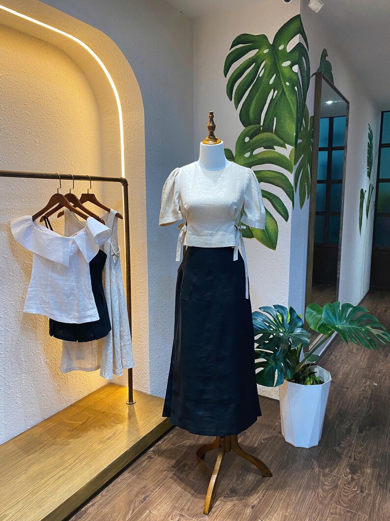 Puede incluir: Una blusa de lino blanca con mangas abullonadas y un lazo en la cintura, combinada con una falda de lino negra. El conjunto se muestra en un maniqu&iacute; en un entorno de tienda.
