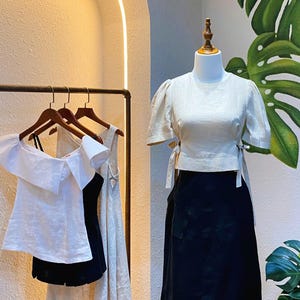 Puede incluir: Tres blusas de lino blanco en perchas, una con cuello de volantes y dos con detalles de lazo. Una blusa de lino blanco con detalles de lazo se muestra en un maniqu&iacute; con una falda negra.