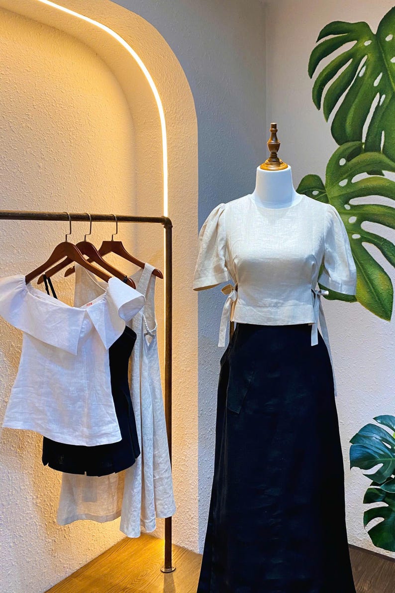 Puede incluir: Tres blusas de lino blanco en perchas, una con cuello de volantes y dos con detalles de lazo. Una blusa de lino blanco con detalles de lazo se muestra en un maniqu&iacute; con una falda negra.