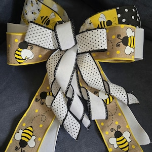 May include: A decorative bow featuring a bee theme. The bow is made of various ribbons, including burlap-style ribbon with yellow polka dots and bee illustrations, white ribbon with black polka dots, and solid white ribbon. The edges are finished with black stitching.