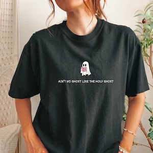 May include: A black short-sleeved t-shirt featuring a small white ghost graphic holding a pink book. Below the ghost, the text reads AIN'T NO GHOST LIKE THE HOLY GHOST in white capital letters. The shirt has a crew neckline and a relaxed fit.