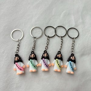 May include: Five keychains featuring small Jesus figurines. Each figurine has black hair and a beard, wearing a white robe and a sash with the text "Jesus ❤ You". The keychains have silver and black metal rings.