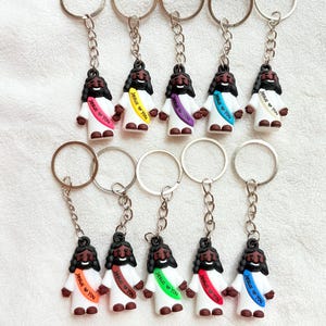 May include: A collection of ten keychains featuring a small, smiling Jesus figure. Each figure has dark hair and a beard, wearing a white robe and a sash with the text "Jesus You" in various colors. The keychains have silver-colored rings and chains.