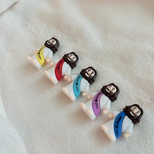 May include: Five small figurines of Jesus with brown hair and beards, wearing white robes and colorful sashes. Each sash has the text "Jesus loves you" in a different color: yellow, red, light blue, purple, and dark blue.