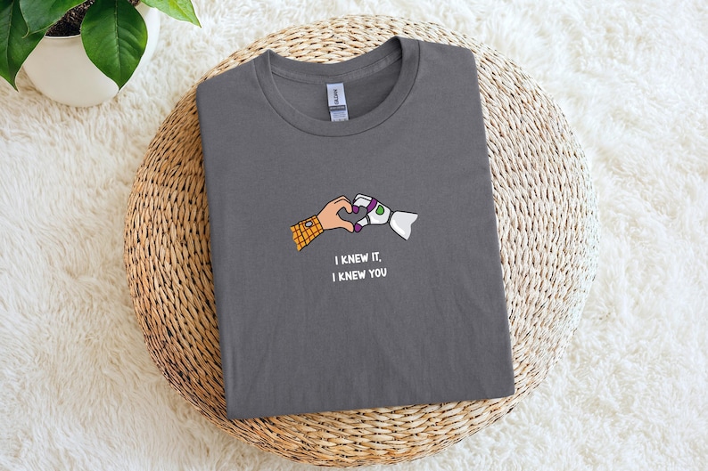 Puede incluir: Una camiseta gris oscuro de cuello redondo con un gr&aacute;fico de dos manos formando un coraz&oacute;n. Una mano lleva una manga de cuadros amarillos y naranjas, mientras que la otra es una mano rob&oacute;tica blanca y verde. El texto "I KNEW IT, I KNEW YOU" aparece debajo.