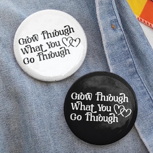 May include: Two round button pins, one white and one black, are pinned to a denim shirt. The pins feature the text "Grow Through What You Go Through" in a stylized font. A rainbow flag is visible in the background. The image also includes the text "BUTTON PIN" and "SHIRTS AVAILABLE".