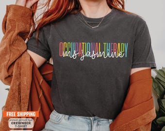 May include: Dark gray crewneck t-shirt with the text "OCCUPATIONAL THERAPY Ms. Jasmine" in colorful letters. The shirt is a garment-dyed style. The image also includes text about free shipping and sweatshirts.