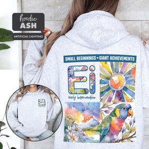 May include: A gray hoodie with a watercolor design on the back. The design features a sun, flowers, a mountain, and a parrot. The text "SMALL BEGINNINGS GIANT ACHIEVEMENTS" and "Ei early intervention" are also included in the design.