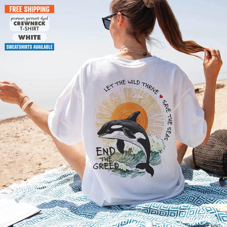 May include: White t-shirt with a graphic of an orca jumping out of the water with a sun and clouds in the background. The text "Let the Wild Thrive" and "Save the Seas" are on the shirt. The text "End the Greed" is below the orca.