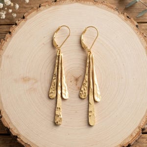 May include: A pair of gold-toned dangle earrings featuring three elongated, hammered-texture metal drops suspended from a lever-back closure. The drops vary slightly in length, creating a tiered effect with a reflective, textured surface finish.