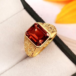 May include: A gold ring featuring a large, rectangular, deep red gemstone set in the center. The band of the ring is crafted with intricate, raised scrollwork patterns that extend along the sides of the setting, surrounding the central stone.