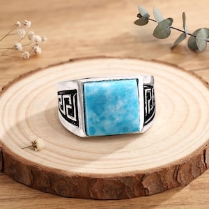 May include: A silver-toned ring featuring a large, square, light blue stone set in the center. The band of the ring is decorated with a black, recessed Greek key pattern on both sides of the stone, providing a high-contrast look to the metal.
