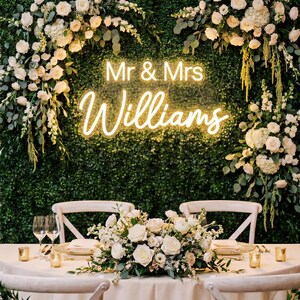 Può includere: Un tavolo da ricevimento di nozze è allestito con rose bianche, candele e caricabatterie dorati. Un'insegna al neon recita "Mr & Mrs Williams" su uno sfondo di verde e fiori bianchi. Due sedie bianche sono di fronte al tavolo.