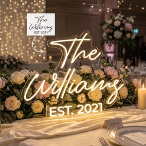 May include: A custom neon sign featuring the text The Williams EST. 2021 in a warm white glow. The sign is mounted on a clear acrylic backing and is positioned behind a floral arrangement of white and pink roses on a table setting.
