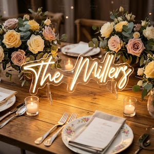 May include: A custom neon sign featuring the text The Millers in a warm white script font, mounted on a clear acrylic backing with two transparent stands, positioned on a wooden table decorated with floral arrangements and lit candles.