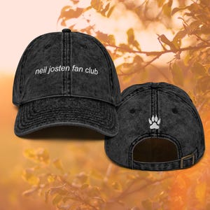 May include: A distressed black baseball cap with the text "neil josten fan club" in white script on the front. The back of the cap features a white paw print and an adjustable strap.