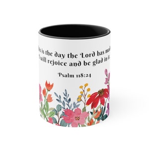 May include: White ceramic mug with a black rim. The mug features a floral design with pink, red, and yellow flowers. The text "This is the day the Lord has made, I will rejoice and be glad in it. Psalm 118:24" is printed on the mug.
