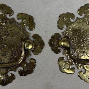 Puede incluir: Dos tiradores de cajón de latón a juego con una placa circular, un intrincado diseño de dragón grabado y un borde festoneado. Cada pieza incluye un asa abatible en forma de U unida al centro, con pequeños orificios para los tornillos.