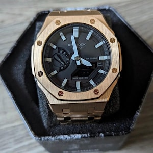 May include: A rose gold-toned G-Shock Casio watch with an octagonal bezel featuring eight visible screws. The watch has a dark grey analog-digital dial with luminous hands, hour markers, a day-of-the-week sub-dial, and a digital display window.