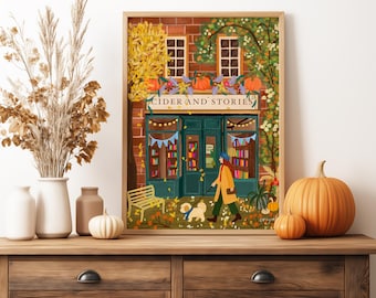 Op de afbeelding: Een ingelijste kunstdruk getiteld CIDER AND STORIES met een bakstenen winkelpui met een boekhandel, een persoon die een kleine witte hond uitlaat, herfstbladeren en pompoenen, geplaatst op een houten ondergrond met droogbloemen.