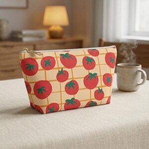 May include: A beige cosmetic bag with a repeating pattern of red tomatoes and green leaves on a yellow grid. The bag has a gold zipper and is sitting on a table.