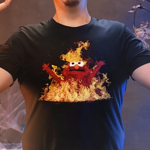 May include: Black t-shirt featuring a cartoon character engulfed in flames. The image is set against a Halloween-themed backdrop with cobwebs, spiders, and string lights. The person in the image has their arms raised.