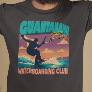 May include: Dark gray t-shirt featuring a graphic of a person waterboarding with the text "GUANTANAMO Bay WATERBOARDING CLUB". The design includes a silhouette of a person on a board, waves, and a sunset.