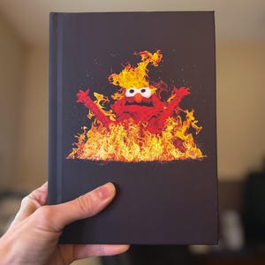 May include: A black book cover featuring a cartoon character engulfed in flames. The character is red with large white eyes and is surrounded by orange and yellow flames. The book is held by a hand.