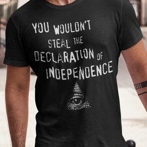 May include: Black t-shirt with white text that reads "YOU WOULDN'T STEAL THE DECLARATION OF INDEPENDENCE." Below the text is a graphic of an eye inside a triangle. The shirt has short sleeves.
