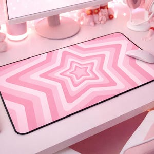 May include: A rectangular desk mat featuring a repeating pattern of concentric stars in varying shades of pink and white. The mat has a black stitched border and is positioned on a white desk surface, with a white computer mouse resting on its right side.