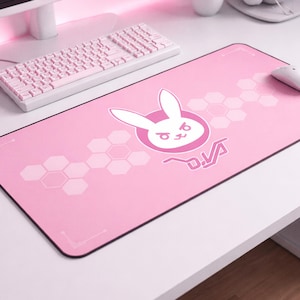 May include: Pink rectangular gaming mouse pad with a white bunny logo and the text "D.Va". The pad has a honeycomb pattern and a white border. A white mouse and keyboard are also visible.