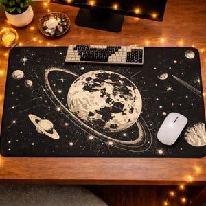 May include: Black rectangular desk mat with a celestial design featuring planets, stars, and rings. A large planet is in the center, with a smaller planet to the left. A white keyboard and mouse are on the mat.