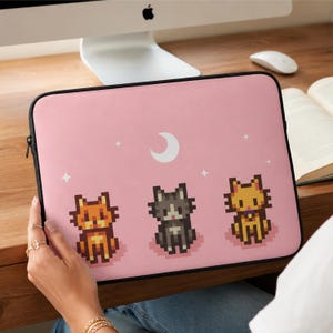 May include: Pink laptop sleeve featuring pixelated cat illustrations. The design includes three cats in orange, gray, and yellow, along with a white crescent moon and stars. The sleeve has a black zipper and is designed to protect a laptop.