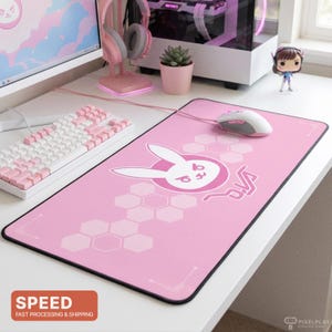 May include: Pink gaming mousepad with a white bunny logo and the word "D.Va" in a stylized font. The mousepad has a black border and a honeycomb pattern. A white and pink keyboard and mouse are also visible.