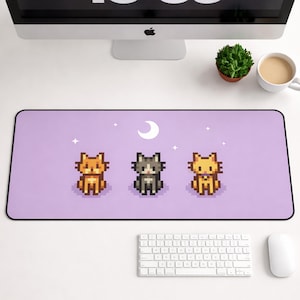 May include: A rectangular lavender desk mat featuring three pixelated cat illustrations in orange, gray, and yellow. A white crescent moon and stars are above the cats. The mat has a black border.