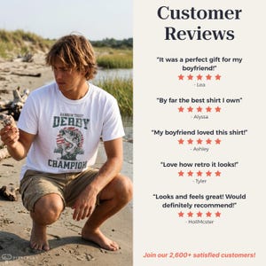 Pu&ograve; includere: T-shirt bianca con grafica di pesce e la scritta "RAINBOW TROUT DERBY CHAMPION". Una persona accovacciata su una spiaggia sabbiosa indossa la maglietta. L'immagine include anche recensioni dei clienti.