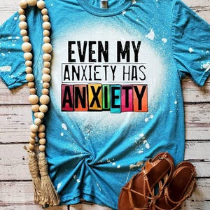 May include: A turquoise t-shirt with the text "EVEN MY ANXIETY HAS ANXIETY" in black and colorful block letters. The shirt has a bleached effect and is paired with brown sandals and a wooden bead necklace. The items are arranged on a wooden surface.