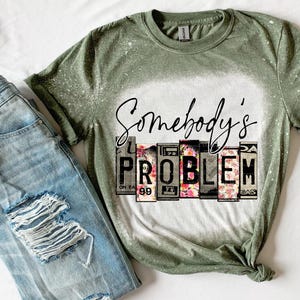 Könnte beinhalten: Olivgrünes T-Shirt mit dem Text "Somebody's Problem" in schwarzer Schrift. Das Wort "PROBLEM" ist in einem Blockbuchstaben-Design mit floralen Akzenten gehalten. Hellblaue Jeans mit Distressed-Details liegen neben dem Shirt.