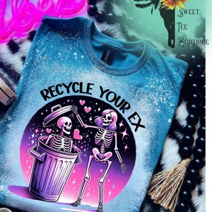 May include: Blue t-shirt with a distressed, bleached effect. The graphic features two skeletons, one in a trash can, and the other dancing, with the text "Recycle Your Ex" in a circular design. The shirt has a crew neck.